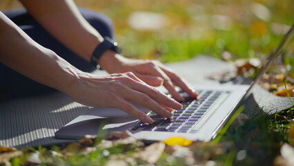 Woman's hands typing on a laptop keyboard while sitting on a yoga mat in a sunny autumn park, embracing flexible remote work and digital nomad lifestyle outdoors