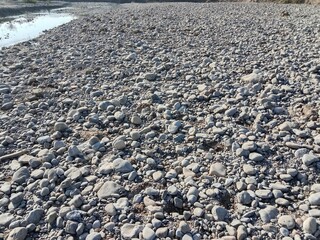Dry riverbed covered with smooth grey pebbles and rounded stones of different sizes creating a natural rocky texture in bright sunlight.