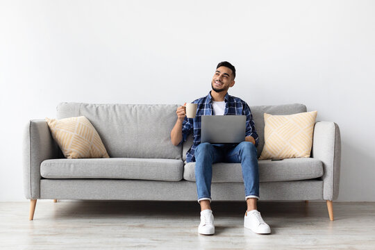 A cheerful man is sitting on a comfortable couch in his living room, sipping coffee and using his laptop. He is enjoying his weekend, feeling relaxed and content.