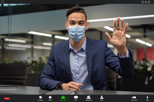 A businessman from the Middle East in a face mask conducts a video call with clients. He looks at the camera and waves, showing engagement in remote business communication.