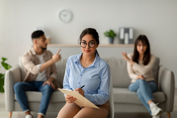 An Arab couple engages in a heated argument on a couch during a therapy session. The female therapist looks at the camera, attempting to manage the situation.