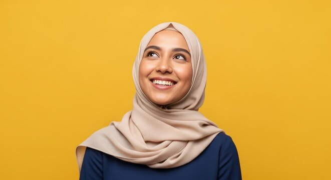Smiling young Muslim woman in a beige hijab looking up with a hopeful and thoughtful expression, standing against a vibrant yellow background, embodying dreams and positive aspirations