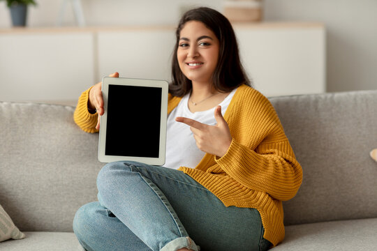 A joyful Arab woman sits on a couch, pointing at a tablet screen. She enjoys shopping online and browsing for gifts at home. The setting is cozy and inviting, perfect for digital purchases.