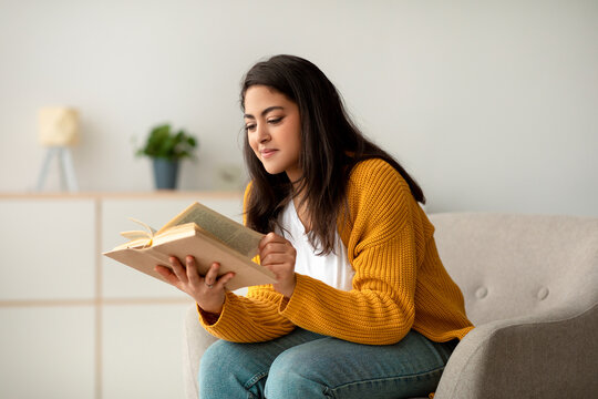 Millennial Arab woman sits in a cozy armchair, reading a book during her break from work. The room is peaceful and inviting, perfect for relaxing on weekends.