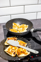 Freshly prepared savory lavash pastries with cottage cheese on a black plate, next to a frying pan on a modern induction stove