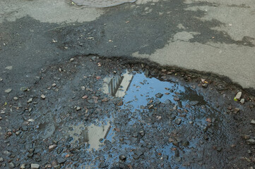 Broken asphalt with a pothole filled with gravel and a puddle reflecting a building.