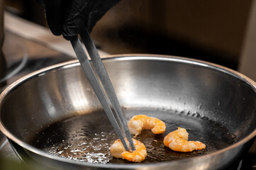 A chef wearing black gloves uses metal tweezers to cook succulent, pink shrimp (prawns) in a hot, stainless steel frying pan with oil.