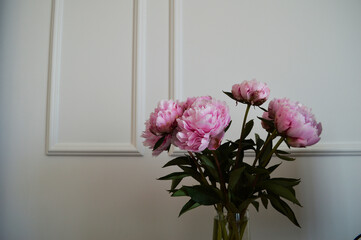 Pink Peonies in a Glass Vase Against White Wall. A bouquet of lush pink peonies with green leaves is arranged in a transparent glass vase set against a classic white wall with molding