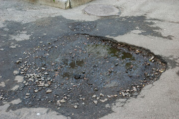 Broken asphalt with a pothole filled with gravel and a puddle reflecting a building.