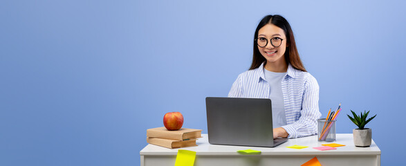 A young woman sits at a clean desk engaged in work on her laptop. Surrounding her are colorful sticky notes, a green plant, books, and a red apple, against a light blue wall.