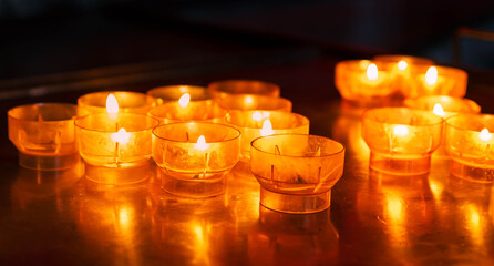 Group of amber-colored church candles burning on a reflective metal surface, creating a warm, peaceful glow in a dark indoor setting