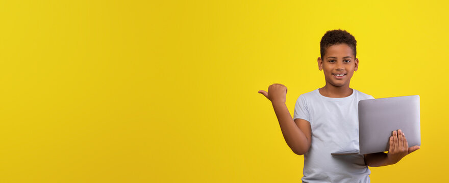 A cheerful boy stands with a laptop in his hand, smiling and pointing to the side. The bright yellow background adds a lively touch to the scene, highlighting his joyful expression. - Powered by Adobe