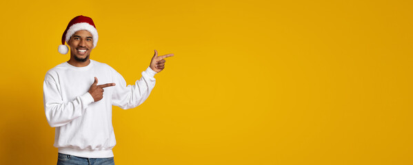 A man with a bright smile is dressed in a white sweater and red Santa hat. He is pointing happily to an unseen object or message on a vibrant yellow background.