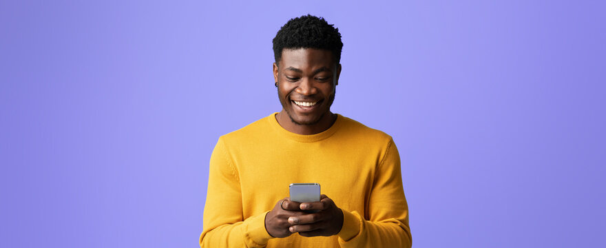A young man with short hair is smiling as he looks at his smartphone. He wears a bright yellow sweater. The bright purple background creates a cheerful and engaging atmosphere. - Powered by Adobe