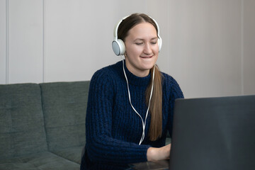 Happy young woman wearing headphones working from home, attending online conference or having a remote lesson, sitting on a sofa and using laptop