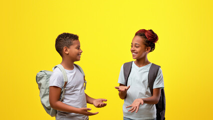 Two kids with backpacks are joyfully talking to each other. They are smiling and standing against a vibrant yellow backdrop, creating a cheerful atmosphere.