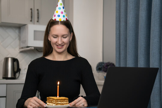 Young woman wearing party hat celebrating birthday online using laptop, sitting in kitchen with piece of cake and lit candle, making video call with friends and family