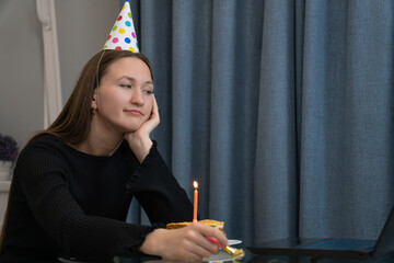 Sad woman with party hat looking at her birthday cake and holding a holiday horn, celebrating alone at home during quarantine, making a video call with her family