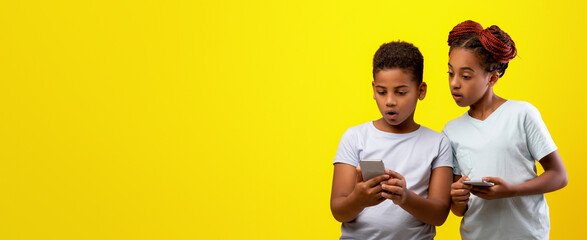 Two kids stand together, engaging with their smartphones. They appear curious and intrigued as they share an experience. The bright yellow backdrop adds a cheerful vibe to the scene.