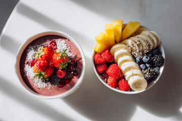 Smoothie bowl paired with sliced banana, berries, and mango on a white table, captured in bright natural light for fresh, colorful breakfast imagery.