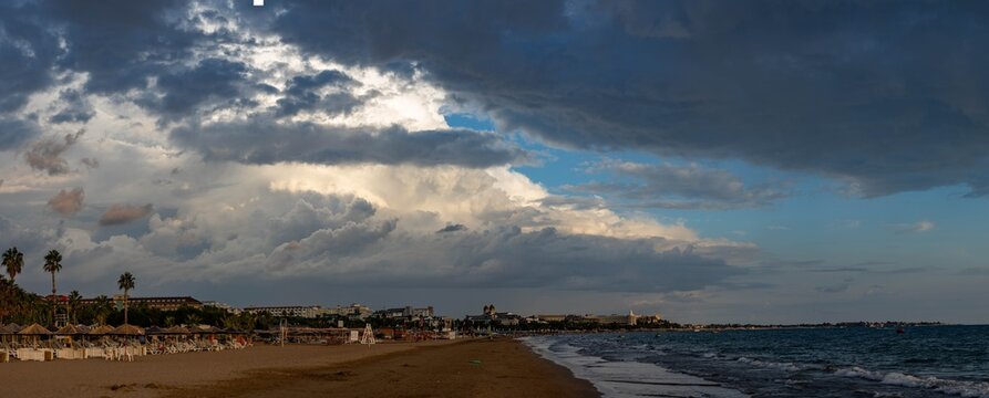 Wide panoramic beach scene with white surf line under changing weather at dusk, showing beach infrastructure and distant hotels under dramatic sky. Evrenseki, Antalya, Turkey.
- Powered by Adobe
