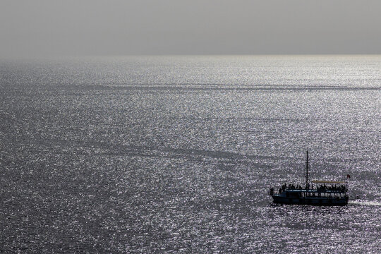 Minimalist view of Mediterranean Sea with sparkling silver surface and pirate ship silhouette in backlight, creating monochromatic blue-gray resort atmosphere. Antalya, Turkey.

