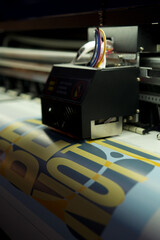 Large format vinyl printer printing yellow and black text on white material with sensor and rollers visible