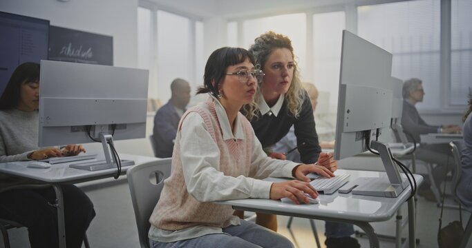 Elegant Female Teacher With Curly Hair Reviews Coding Projects With Adult Students in Bright Modern IT Classroom. Concept Digital Transformation in Education, Support, and Teamwork in Learning.