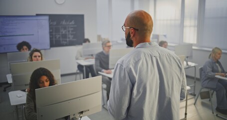 Professional Male Instructor Stands in Front of Adult Students in Computer Lab, Explaining Coding Principles. Concept Continuous Education, Digital Skills, and Modern Learning Environment. Back View.