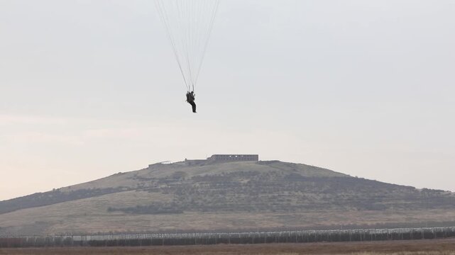 Aerial high-angle shot of a lone skydiver safely descending under a bright white parachute. The skydiver is landing on a vast, dry, military training field near a distant village.