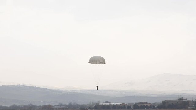 Aerial high-angle shot of a lone skydiver safely descending under a bright white parachute. The skydiver is landing on a vast, dry, military training field near a distant village.