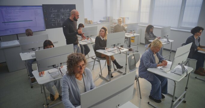 Multiethnic Group of Mature Students Study Together in Bright Modern Computer Classroom While Adult Teacher Walks Around Guiding, Assisting. Concept Lifelong Learning, Digital Inclusion. High Angle.