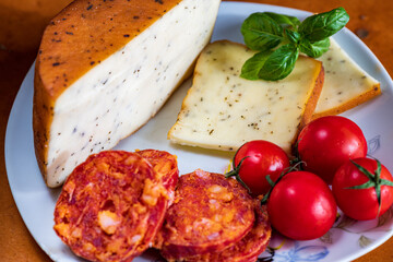 Sliced kulen and semi-hard cheese arranged neatly on a plate, garnished with fresh basil leaves. The vibrant colors of the red cured sausage, creamy-yellow cheese, and green basil create a visually ap