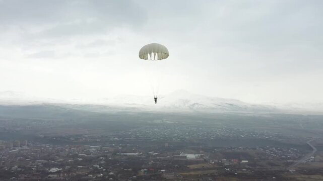 Aerial high-angle shot of a lone skydiver safely descending under a bright white parachute. The skydiver is landing on a vast, dry, military training field near a distant village.