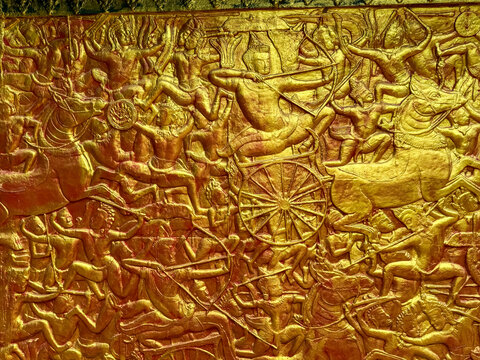 A magnificent and highly detailed golden bas-relief carving on a temple wall in Phnom Penh, Cambodia, depicts an epic mythological battle scene from the Reamker with warriors and chariots.