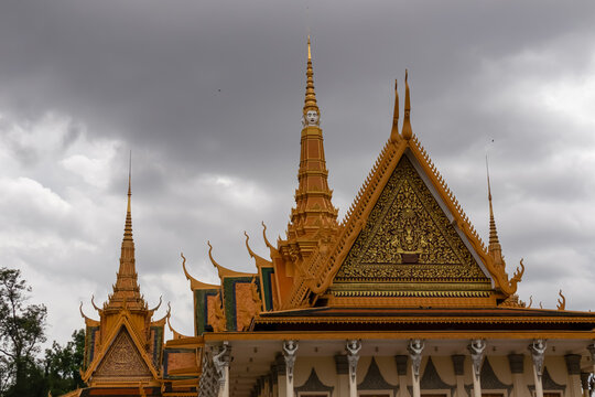 The intricate golden spires and ornate gables of Silver Pagoda, a classic example of Khmer architecture, rise majestically against a dramatic, dark, and cloudy sky at the Royal Palace in Phnom Penh.
