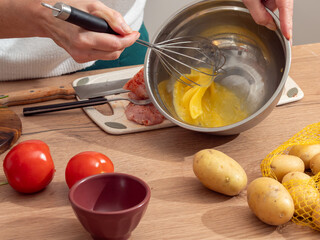 Close-up of a woman whisking eggs in a metal bowl while preparing a meal. Fresh vegetables and raw meat on the table. Authentic home cooking atmosphere.