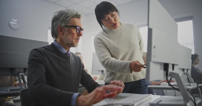 Elegant Grey Haired Man in Glasses Works on Computer While Young Teacher Leans Over, Pointing Screen and Explaining Project Details. Adult Learning, Digital Mentorship, Intergenerational Education.