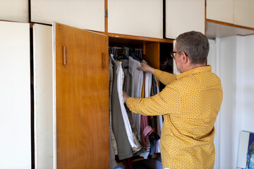 Middle-aged man in a yellow polka-dot shirt opening a wooden closet and choosing clothes, preparing for the day.