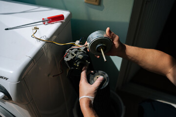 Close-up cropped shot of unrecognizable technician wearing safety gloves examines components of disassembled washing machine, focusing on detached motor while troubleshooting malfunction.