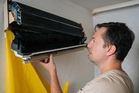 Thoughtful technician disassembling and repairing wall-mounted air conditioner unit, using drainage bag to collect water while working on electrical components. Concept of air condition maintenance