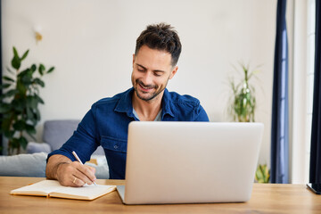 Man at home writing in notebook using laptop sitting at table in living room