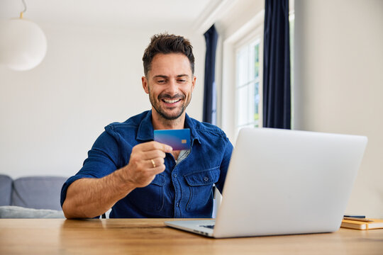 Smiling man sitting at home paying bills online with credit card and laptop