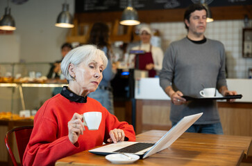 Fototapeta premium Elderly woman sitting at table, using laptop and enjoing cup of tea in coffee house