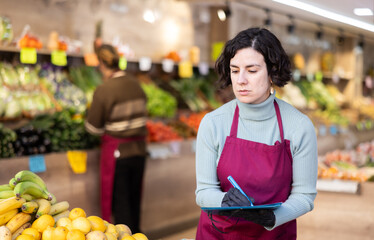 Adult female salesperson taking inventory with list in vegetable store