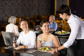 Young male waiter in uniform serves ordered dish to two women in restaurant..