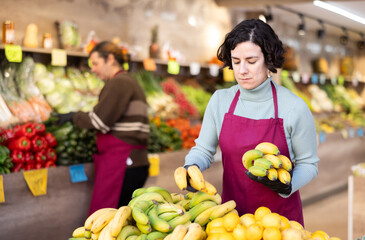 Adult woman seller in apron puts bananas on display in vegetable shop
