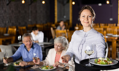 Portrait of polite hospitable waitress holding serving tray with delicious salad and glass of wine meeting guests in cozy restaurant hall