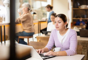 Smiling young woman using laptop and drinking coffee in modern cafe