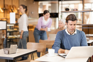 Positive European middle-aged man drinking and enjoying coffee while working on laptop in cafe. Freelance and remote work